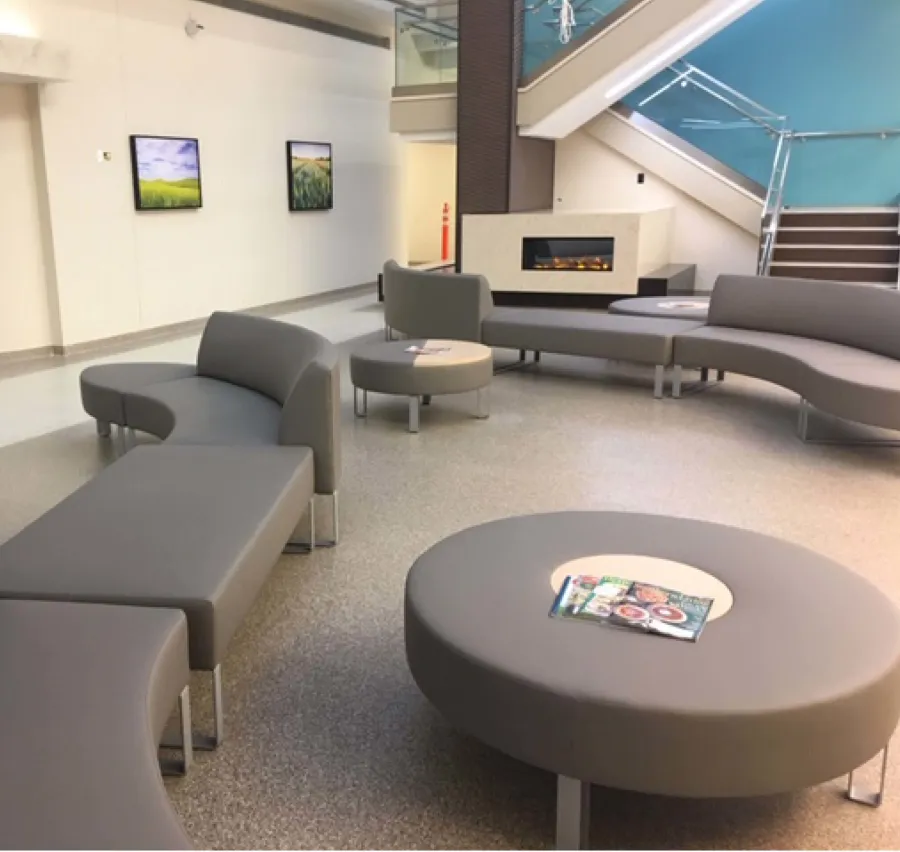 Modern lobby area with curved gray couches, round tables, a fireplace, and stairwell with glass railing.