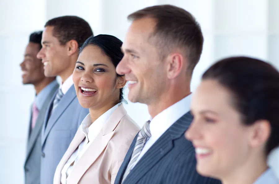 Diverse group of smiling business professionals standing in a row looking confident in office attire.