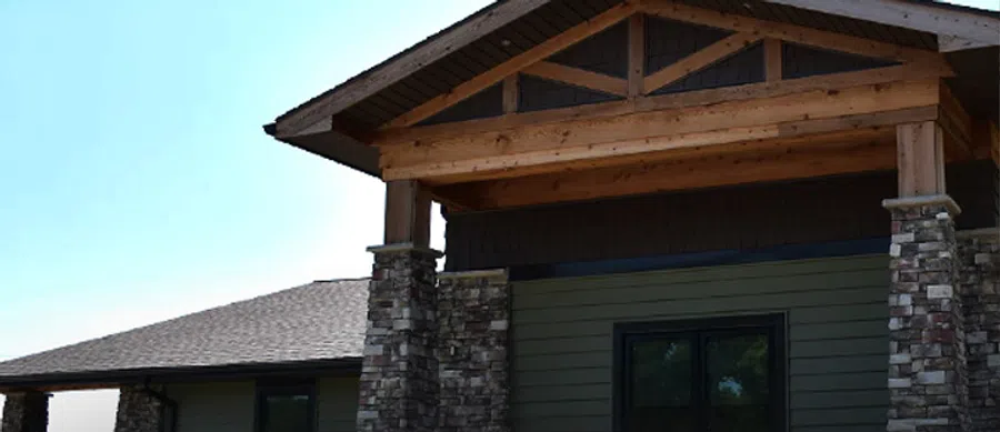 Modern house entrance with stone pillars, wooden beams, and a dark gray roof under clear blue sky.