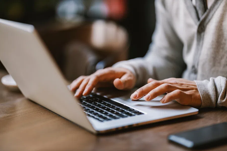 Hands typing on a laptop keyboard on a wooden table with a smartphone nearby in soft natural light