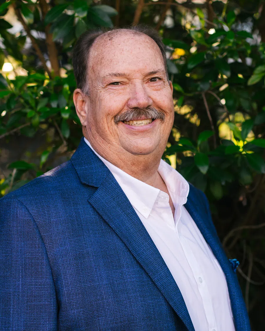 Smiling middle-aged man with mustache wearing a blue blazer and white shirt standing outdoors with green foliage.