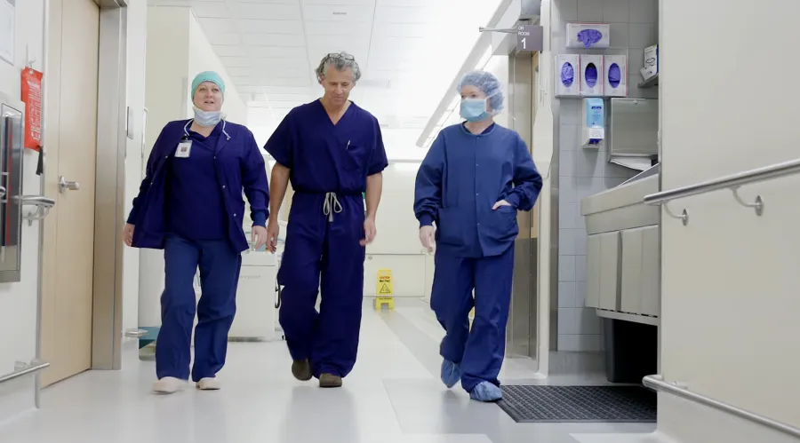 Three medical professionals in scrubs walking down a hospital corridor with equipment and safety signage.