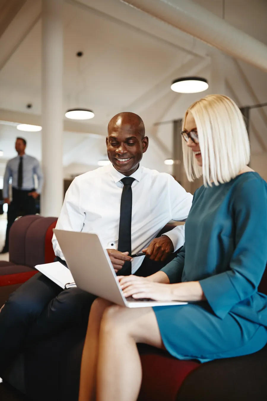 Two business professionals smiling while working on a laptop in a modern office lounge space.