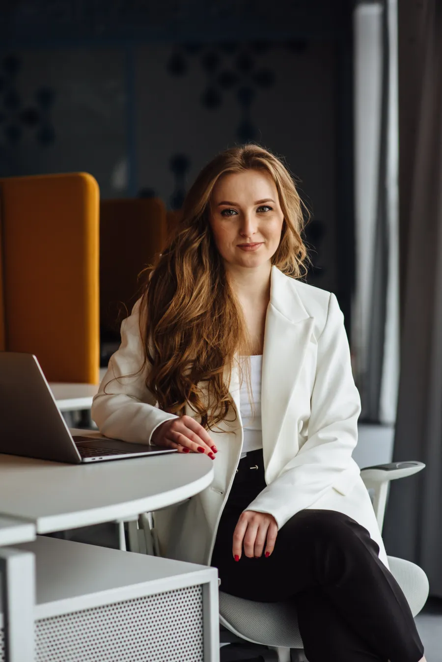 Confident woman with long hair wearing a white blazer seated at office desk with a laptop.