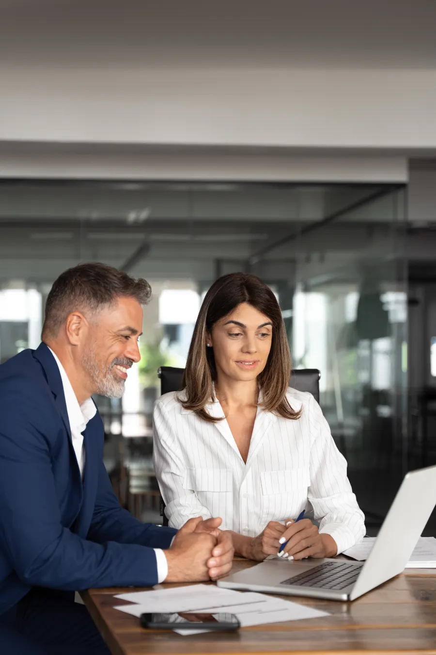 Man and woman collaborating on laptop in modern office with documents on wooden table