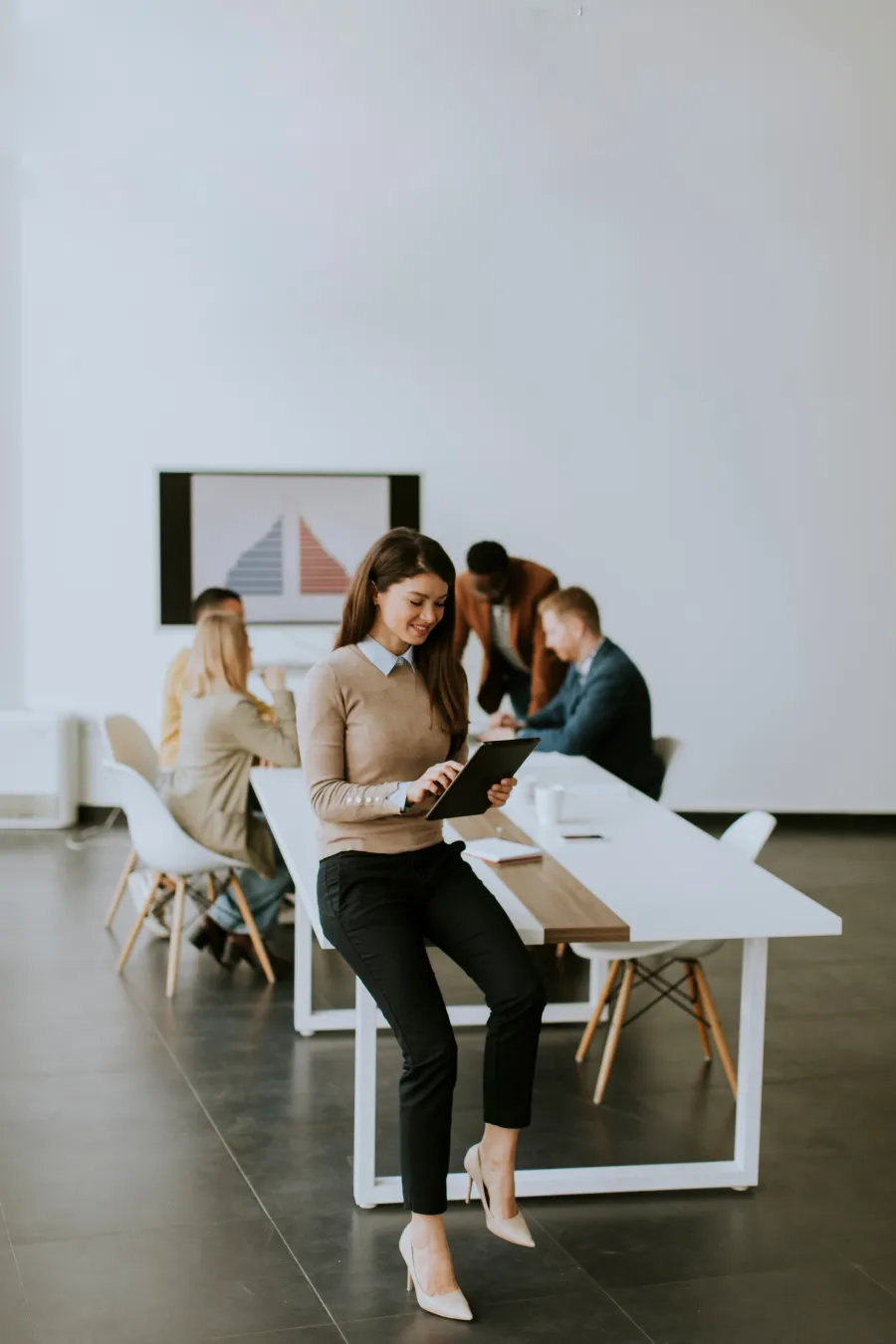 Young businesswoman using tablet while leaning on table in modern office with colleagues meeting in background