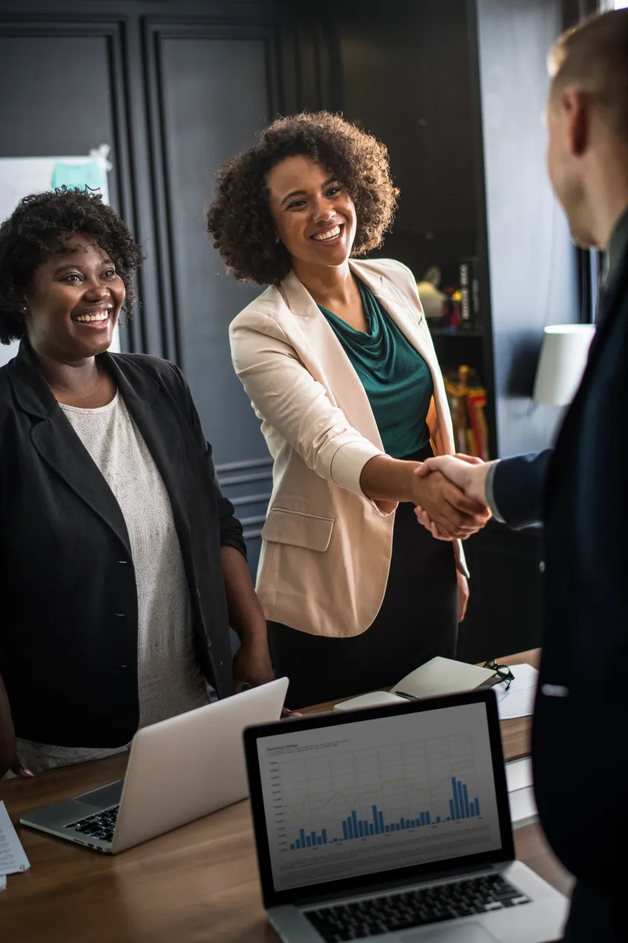Two women smiling and shaking hands with a man in a business meeting with laptops and charts