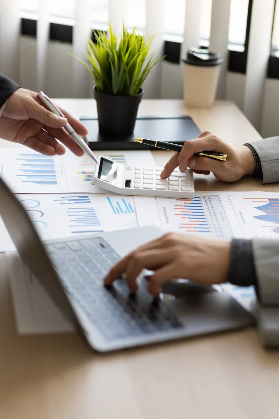 Two professionals analyzing financial charts using a calculator and laptop at a desk with a plant and coffee cup.