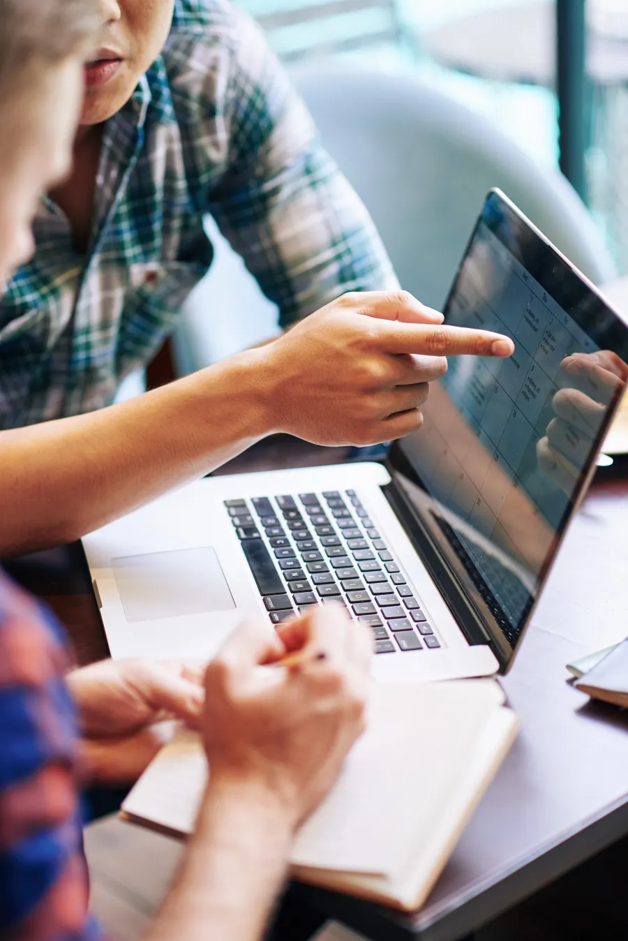 Two people collaborating on a project using a laptop and notebook in a modern workspace.
