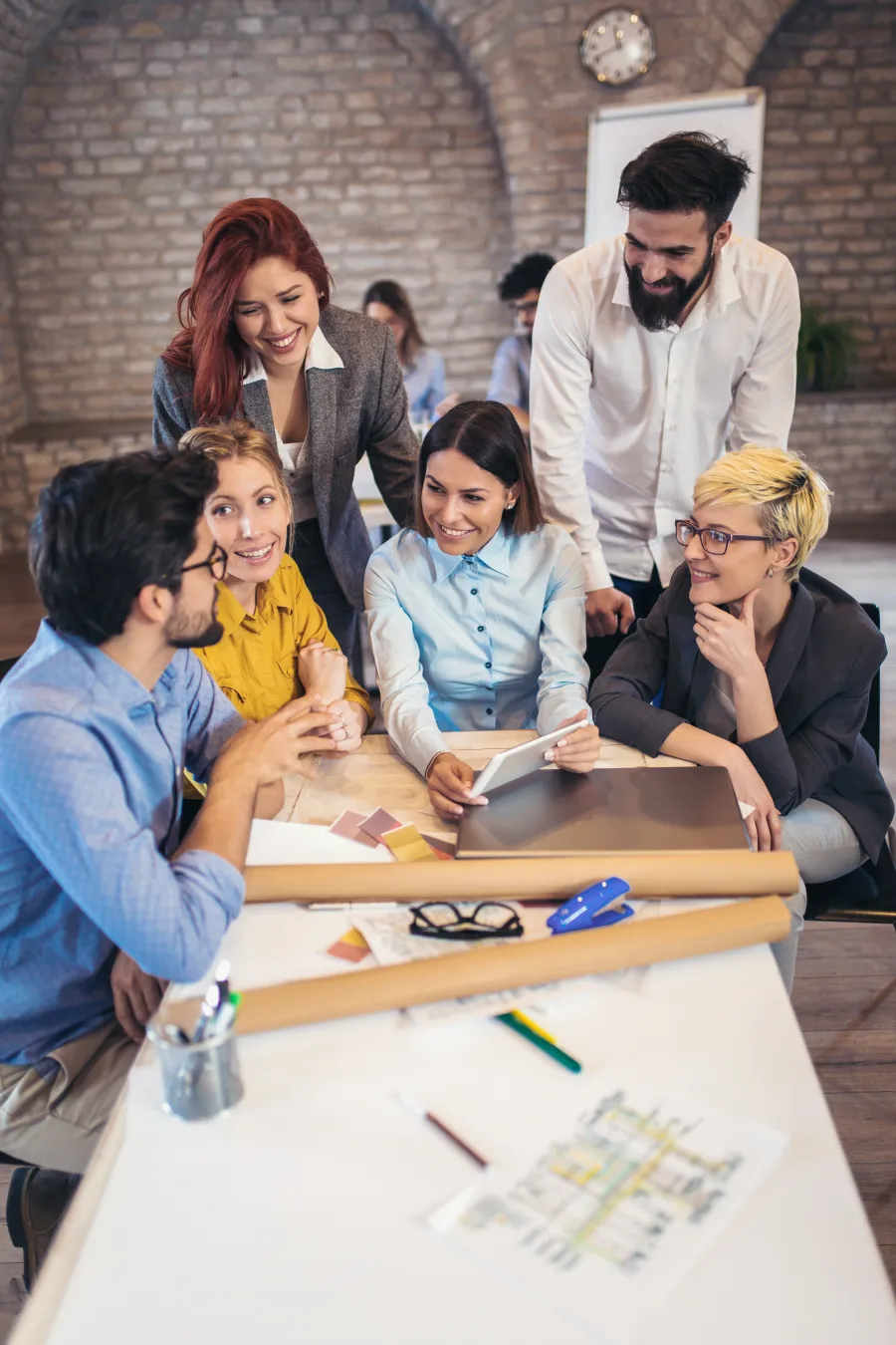 Team of six young professionals collaborating happily around a table with design plans in a modern office.