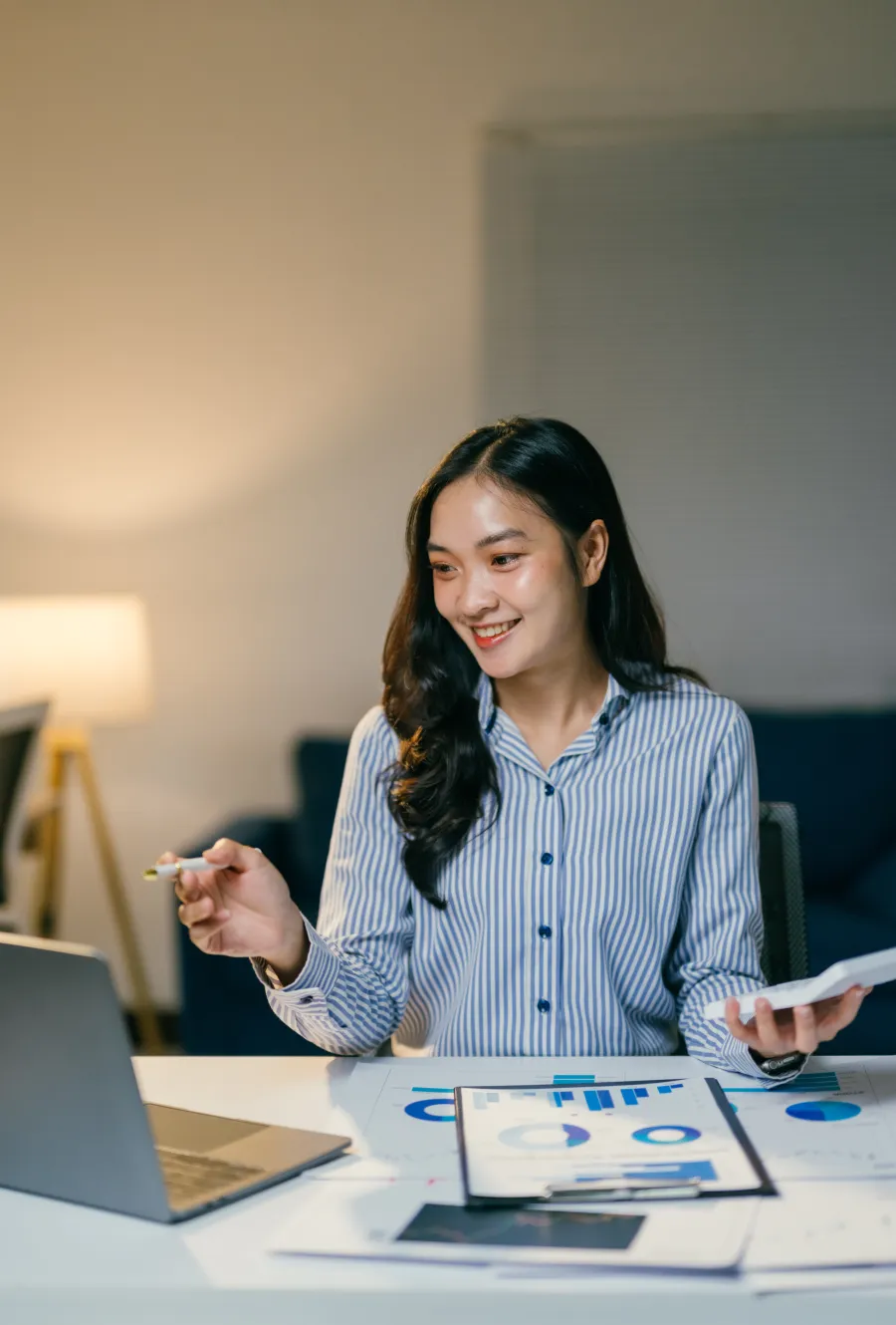 Smiling woman presenting data on a laptop while holding a tablet in a modern workspace.
