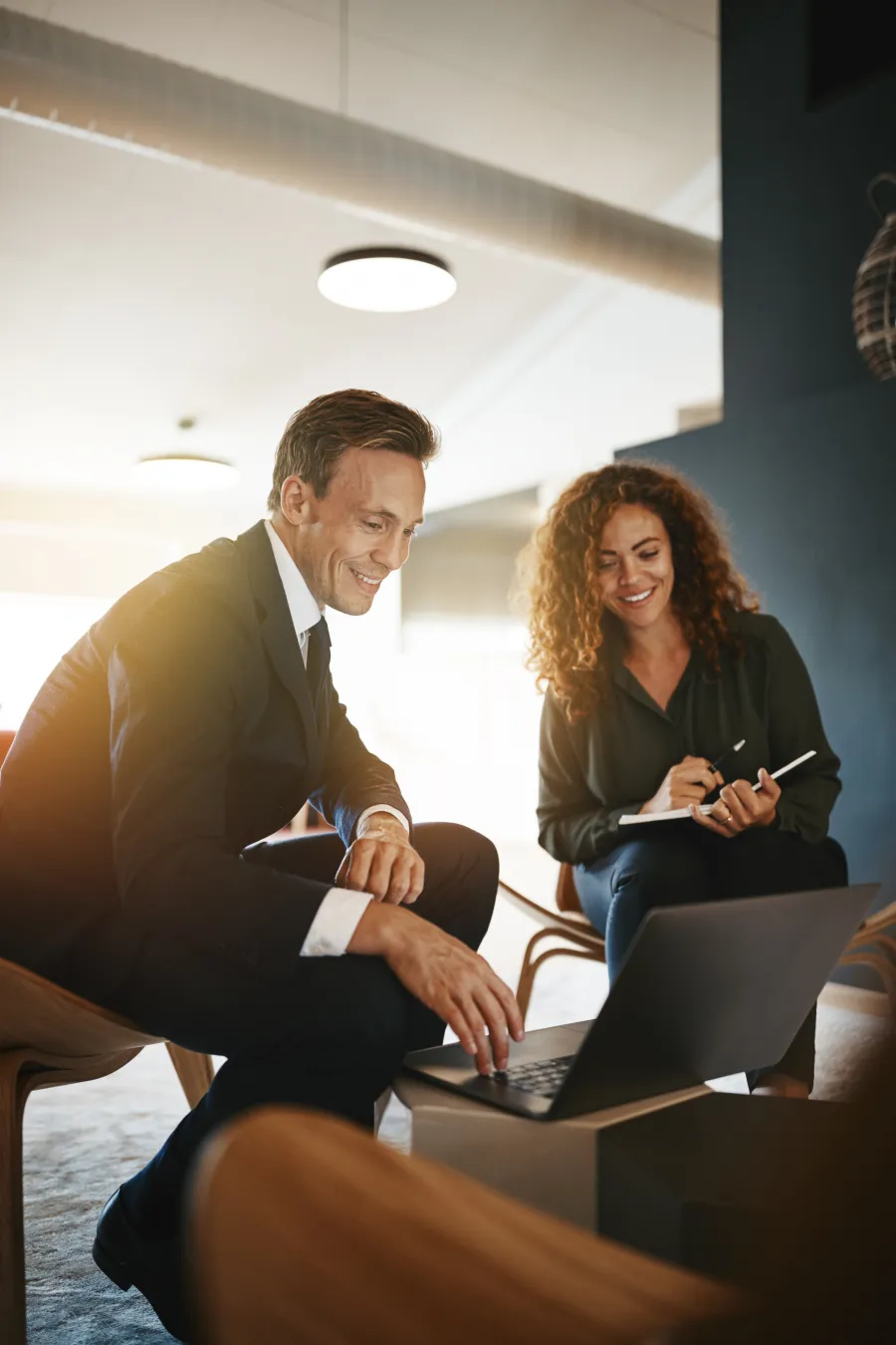 Businessman and businesswoman collaborating on laptop in modern office setting, smiling and taking notes.