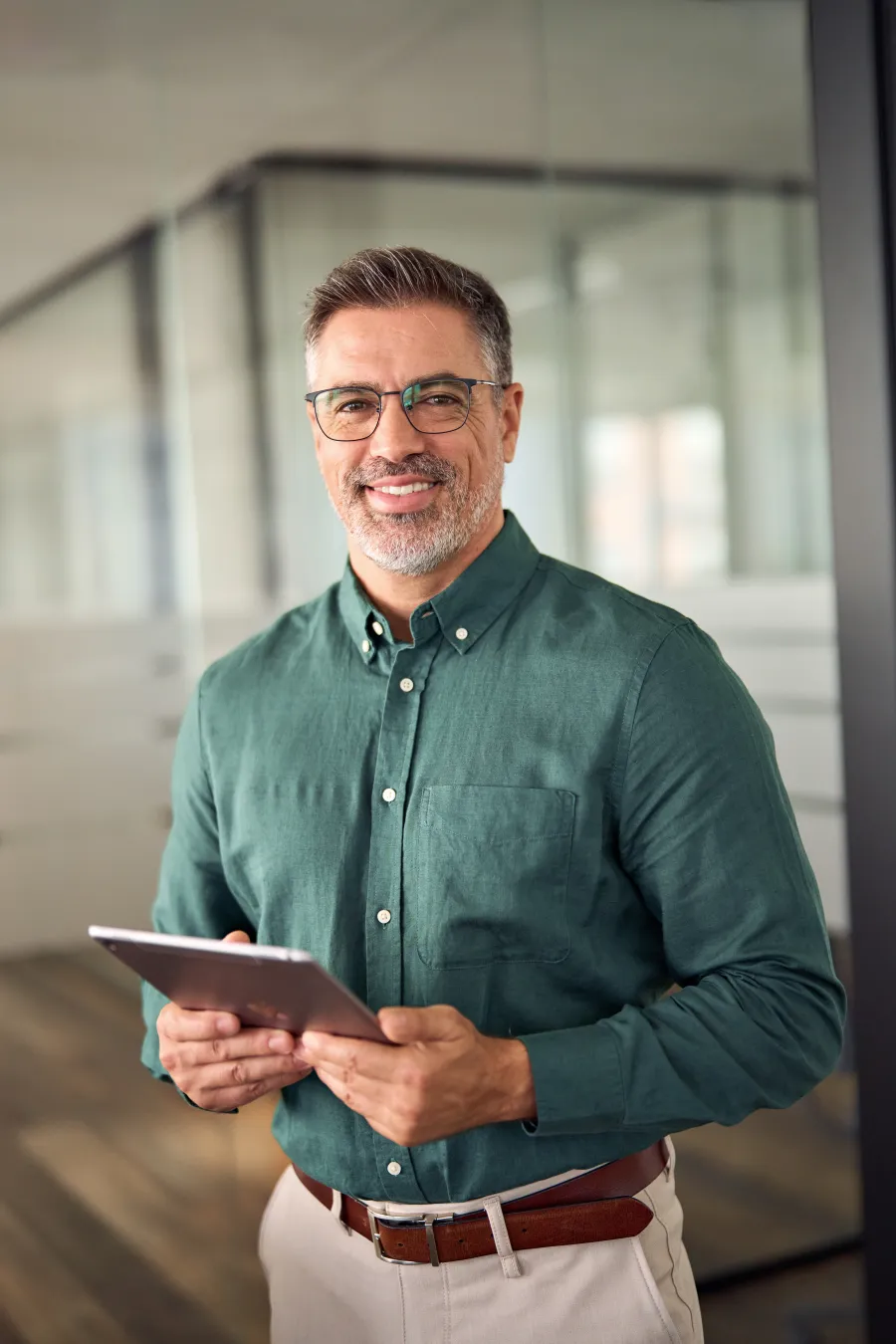 Middle-aged man with glasses smiling and holding a tablet in a modern office setting.