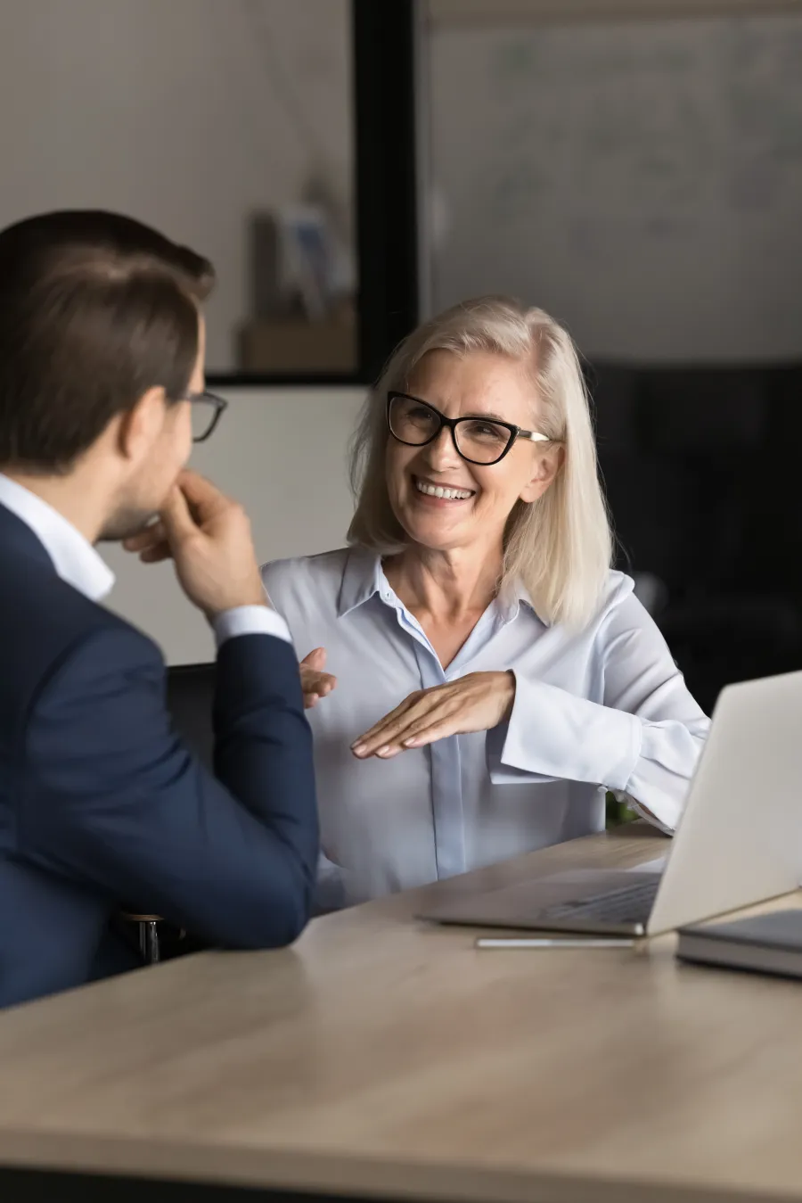 Smiling senior woman with glasses discussing work with a male colleague at office desk with laptop.