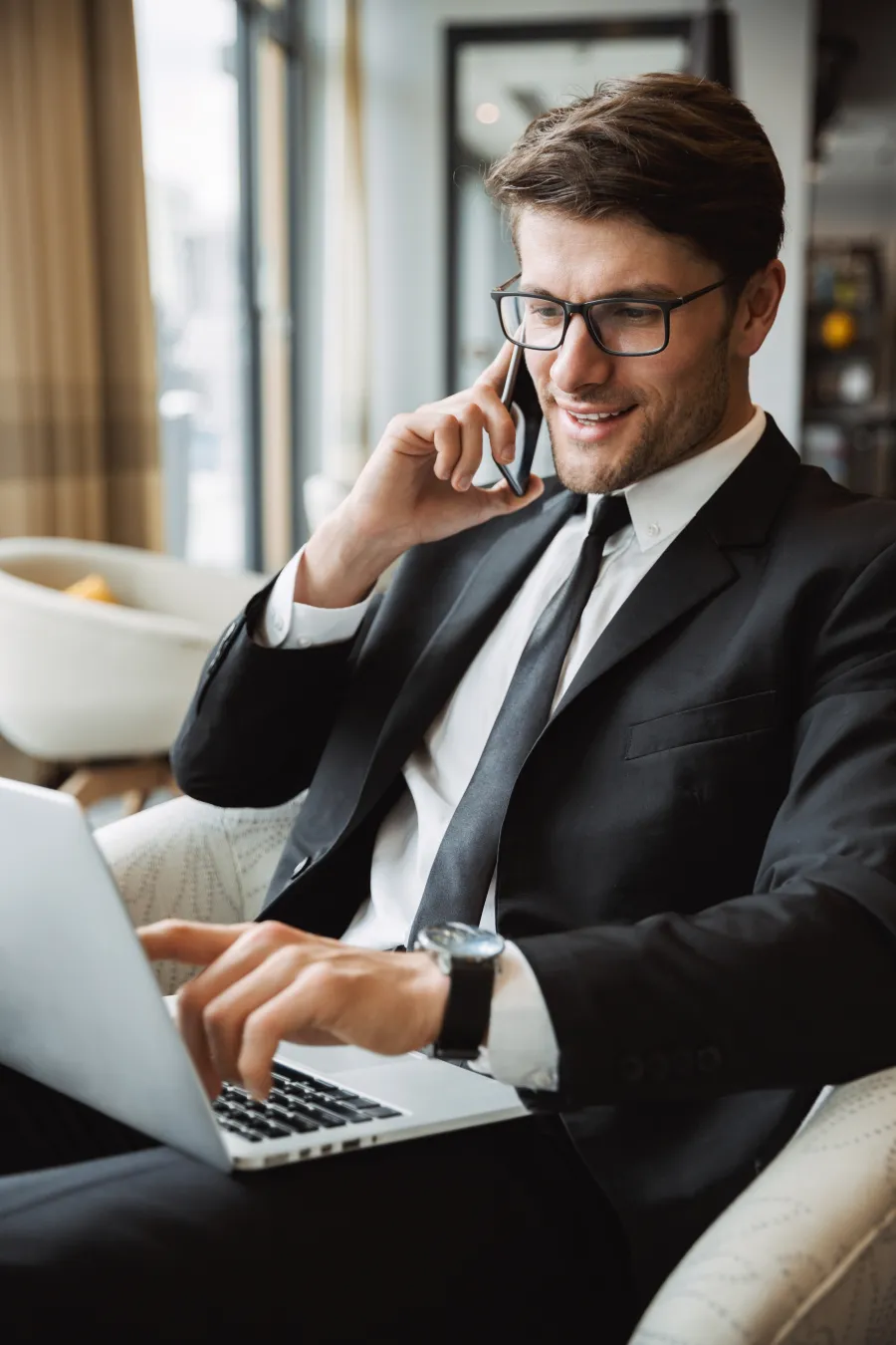 Young businessman in glasses talking on phone and working on laptop in a modern office setting.