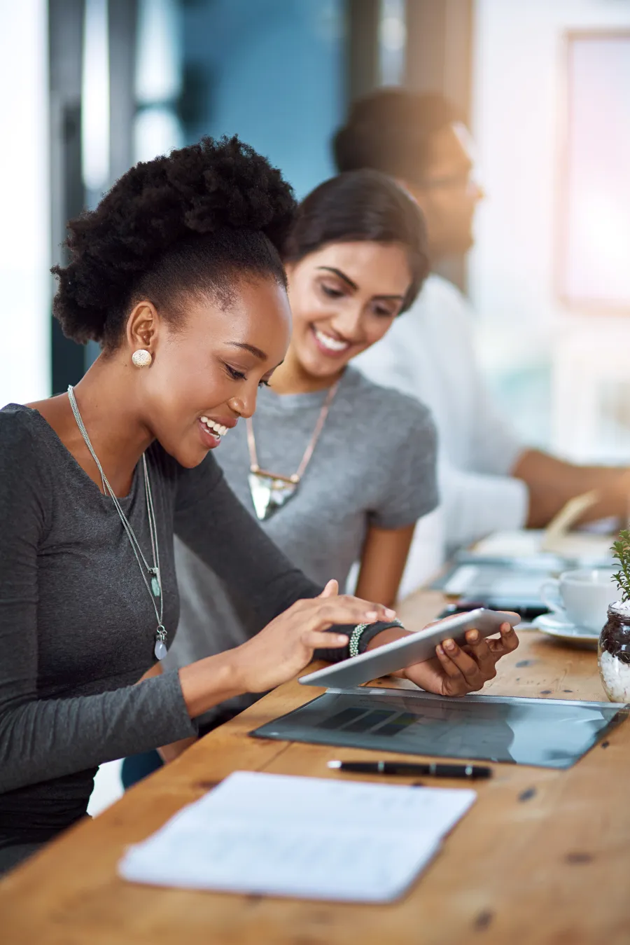 Two women smiling and looking at a digital tablet while seated at a wooden table in a modern office.