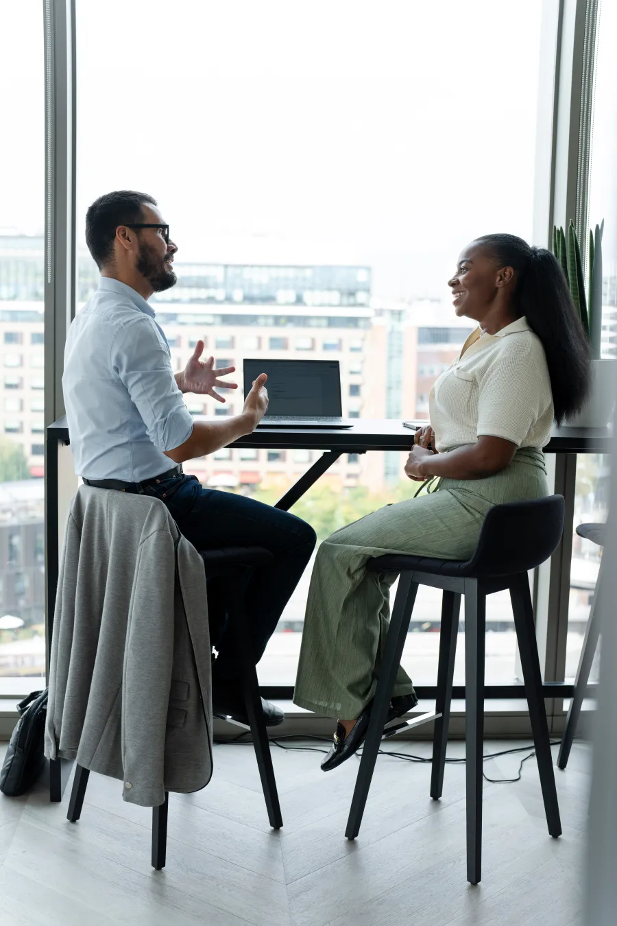 Man and woman having a discussion at a high table with a laptop in a modern office with city views.