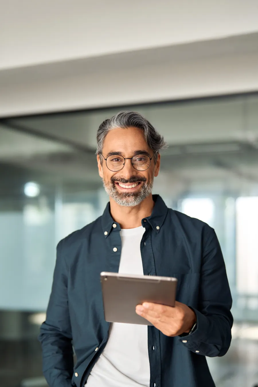 Smiling mature man with gray hair and glasses holding a tablet in a bright modern office space.