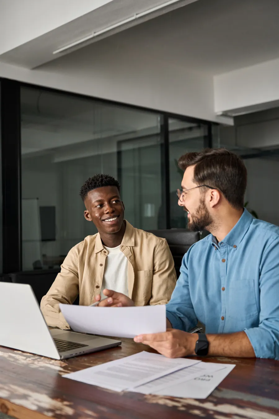 Two coworkers discussing documents and smiling at a table with laptop in a modern office setting.