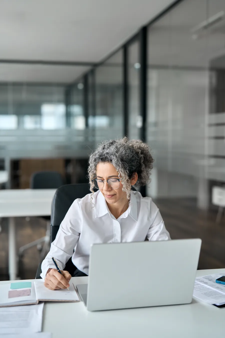 Professional woman with curly hair and glasses working at a laptop and taking notes in a modern office.
