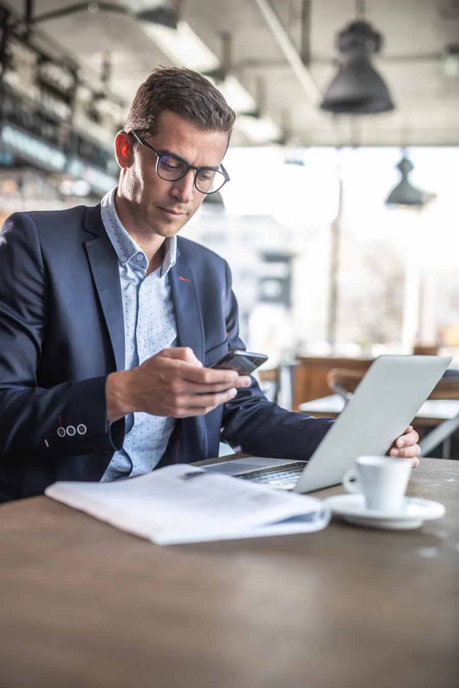 Businessman in glasses using smartphone and laptop at a cafe table with coffee and documents.