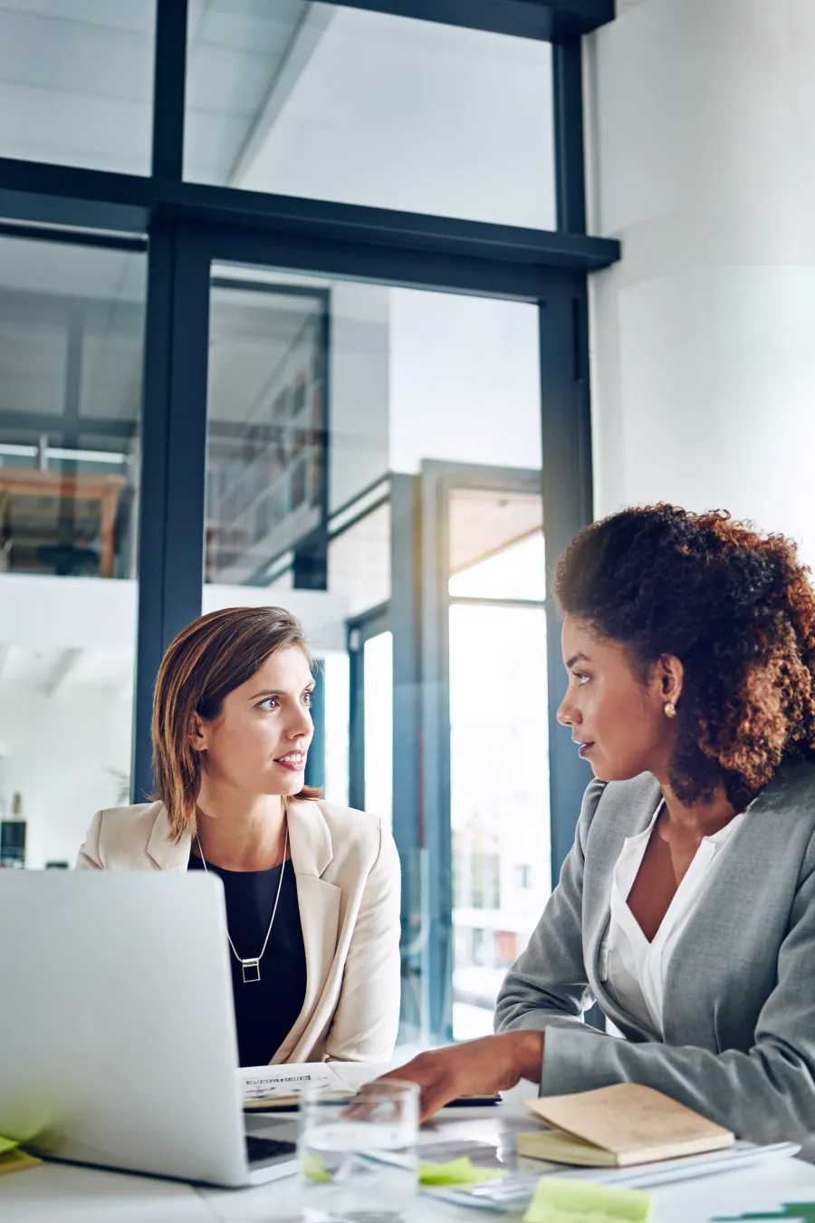 Two businesswomen in suits having a discussion in a modern office with laptop and documents on the table.