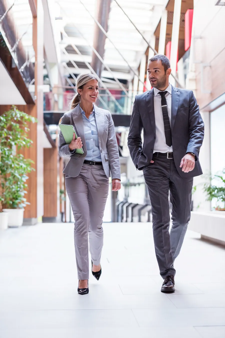 Two business professionals in suits walking and conversing in a modern office hallway with plants.