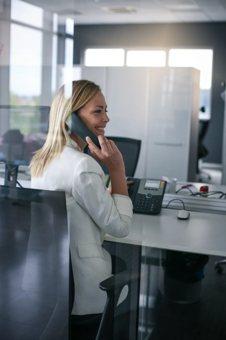 Smiling woman in white blazer talking on office phone at her modern workstation during daytime.