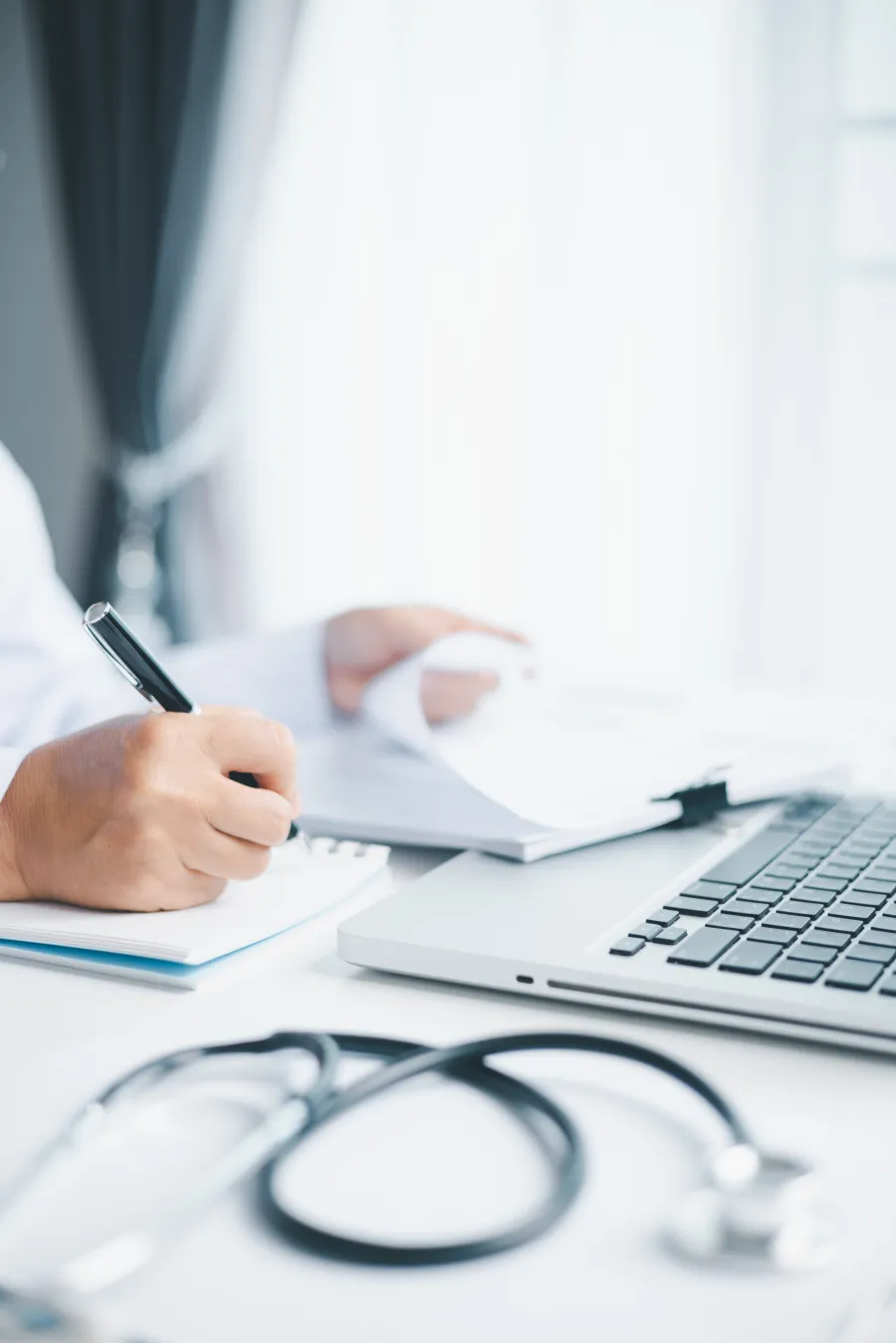 Doctor reviewing medical documents and taking notes next to a laptop with a stethoscope on the desk.
