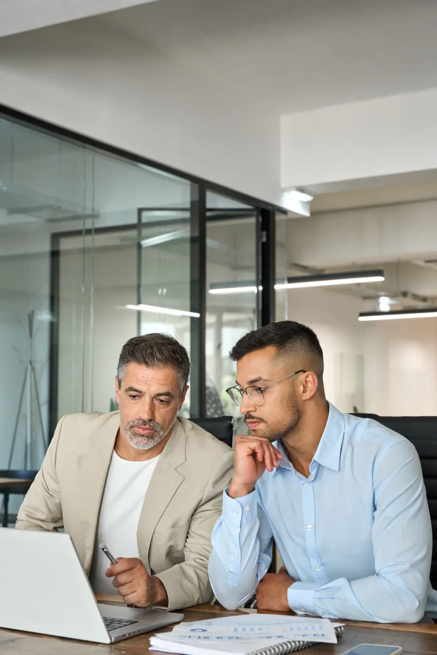 Two businessmen studying data on a laptop and documents in a modern office meeting room.