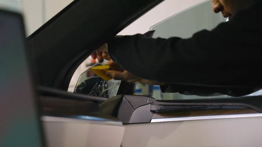 Person installing window tint on car's side door glass using a yellow tool inside vehicle.