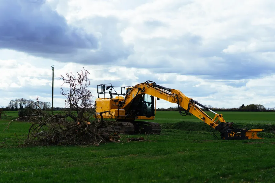 Yellow excavator clearing fallen tree branches on green field under a cloudy sky during daytime