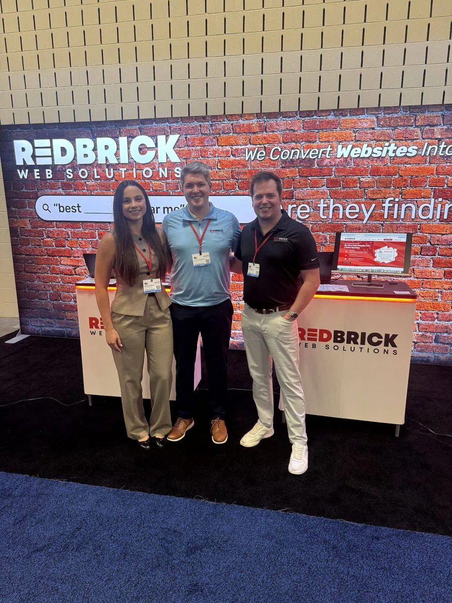 Three professionals standing in front of RedBrick Web Solutions booth with a brick wall backdrop and computer display.