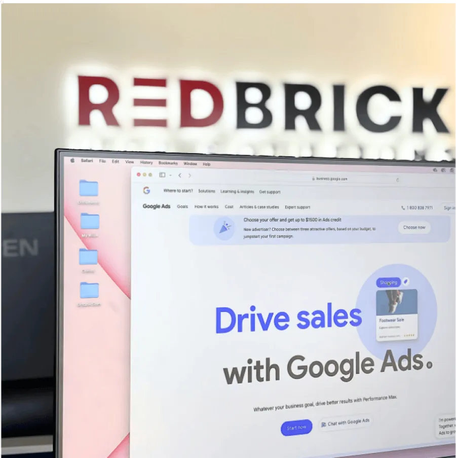 Computer screen showing Google Ads page with RedBrick logo blurred in the background on office wall.