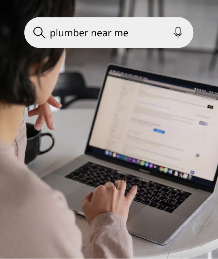 Person using laptop to search online for plumber near me, showing search bar with text and microphone icon.
