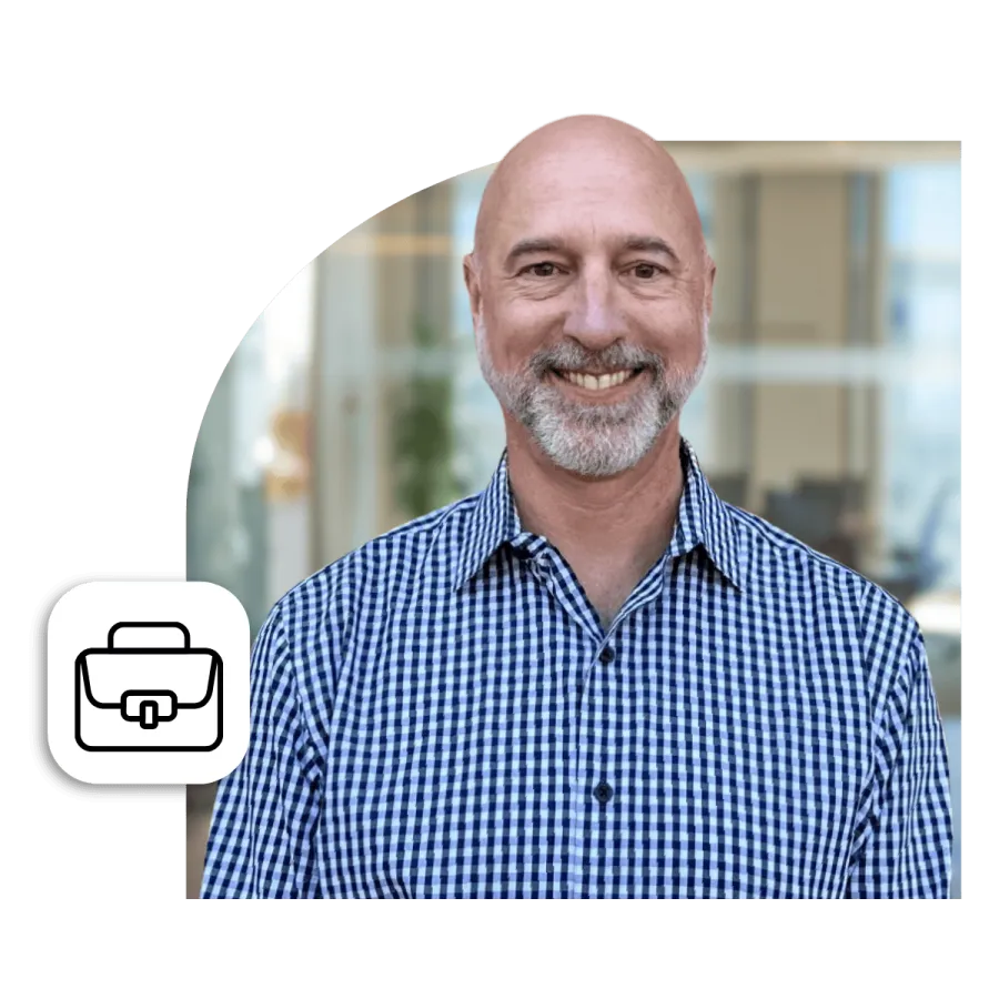 Smiling bald man with gray beard wearing a blue checkered shirt in a modern office setting