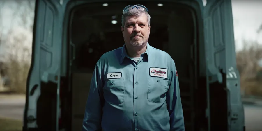 Middle-aged male technician named Chris in a blue work uniform standing in front of an open van door outdoors