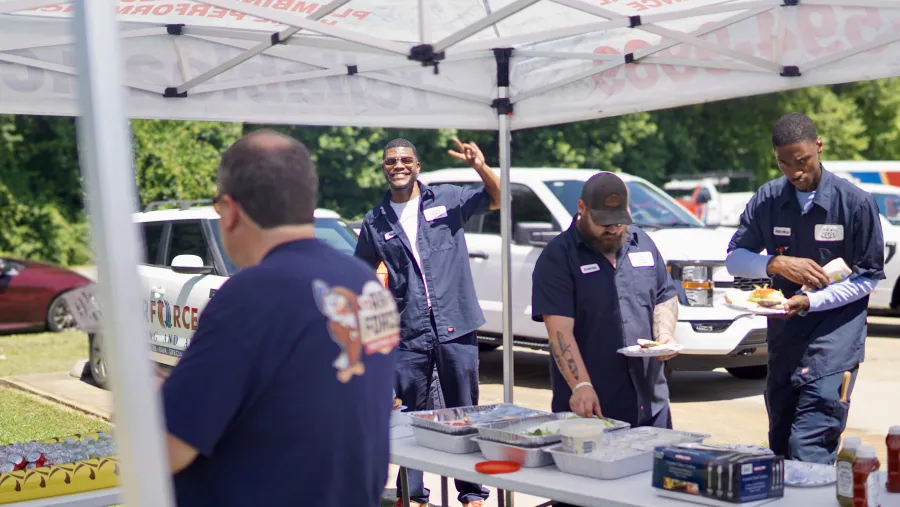 Workers in blue uniforms enjoying a meal under a canopy tent at an outdoor company event with vehicles in background.
