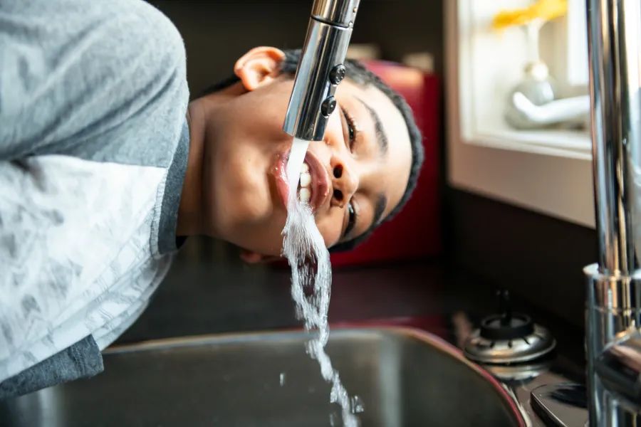Child drinking water directly from a kitchen sink faucet with a stainless steel finish under natural light.