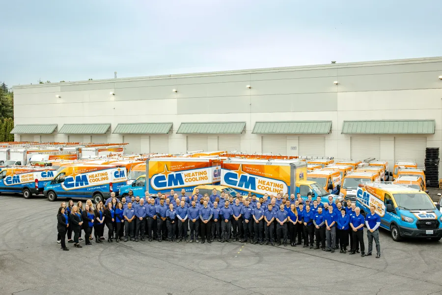 Large group of CM Heating Cooling employees posing in front of company service vans outside warehouse.