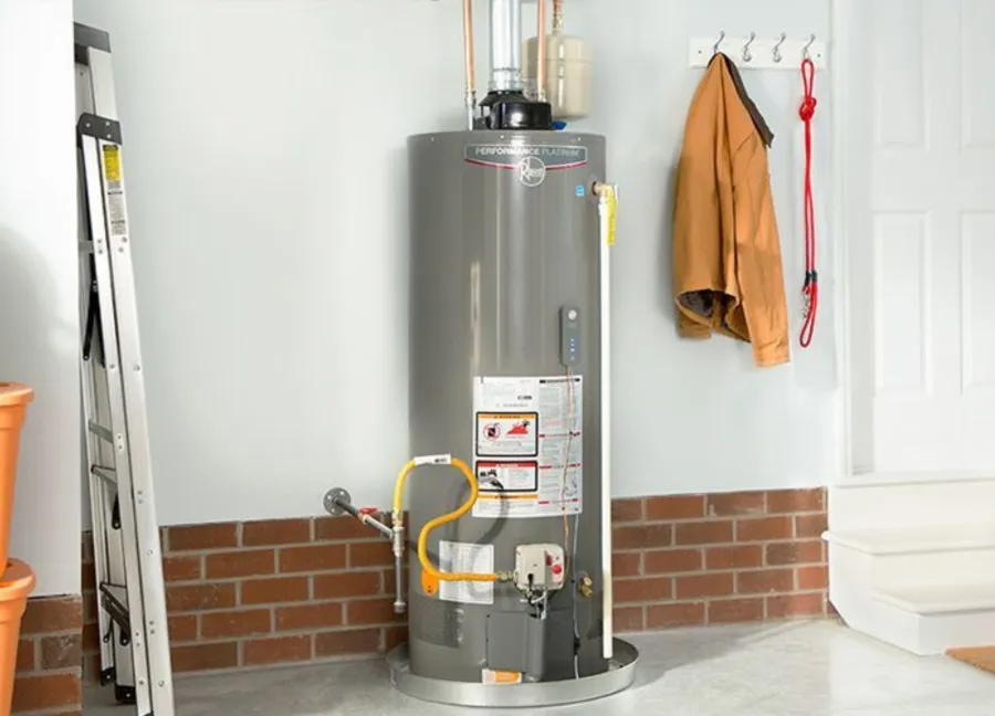 Gray water heater tank in a clean utility room next to a ladder and hanging jacket on a brick half-wall.