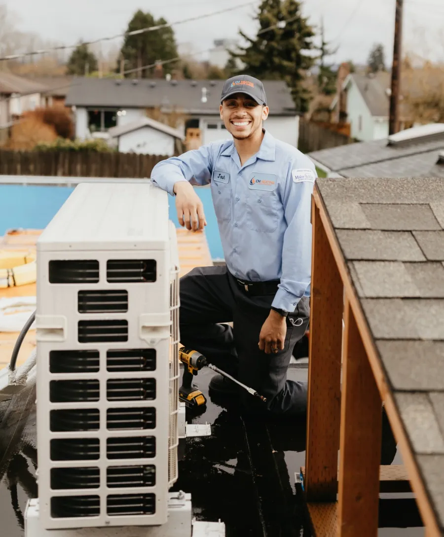 Technician in uniform kneeling on rooftop next to HVAC unit with tools, smiling at camera in residential area.
