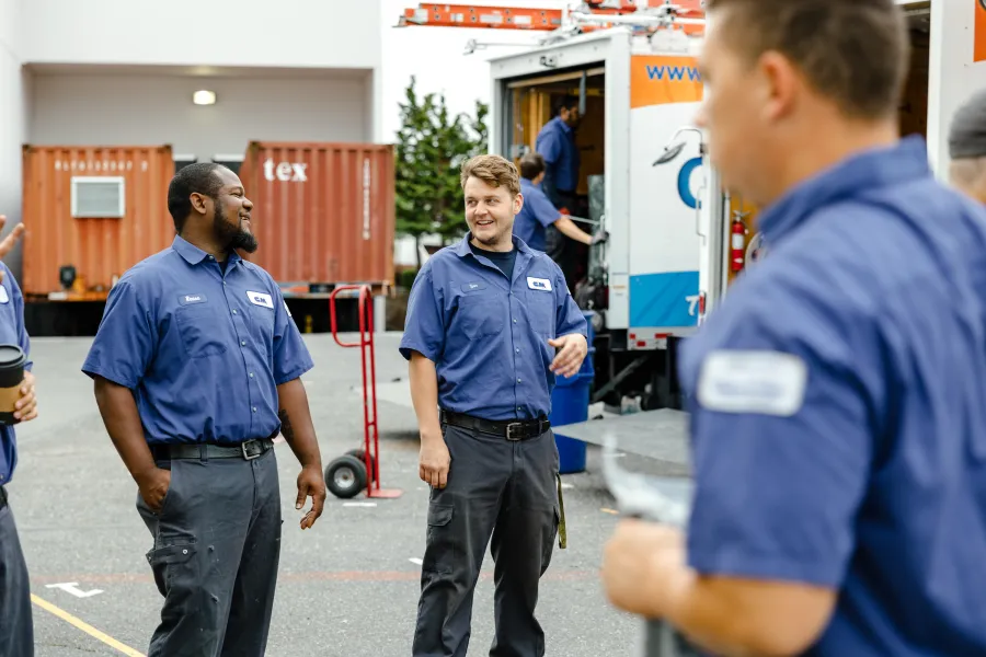 Group of uniformed technicians smiling and talking outside near work trucks and equipment on a sunny day