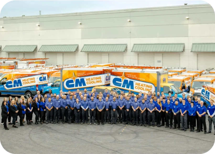 Large group of CM Heating Cooling employees posing in front of branded service trucks outside a warehouse