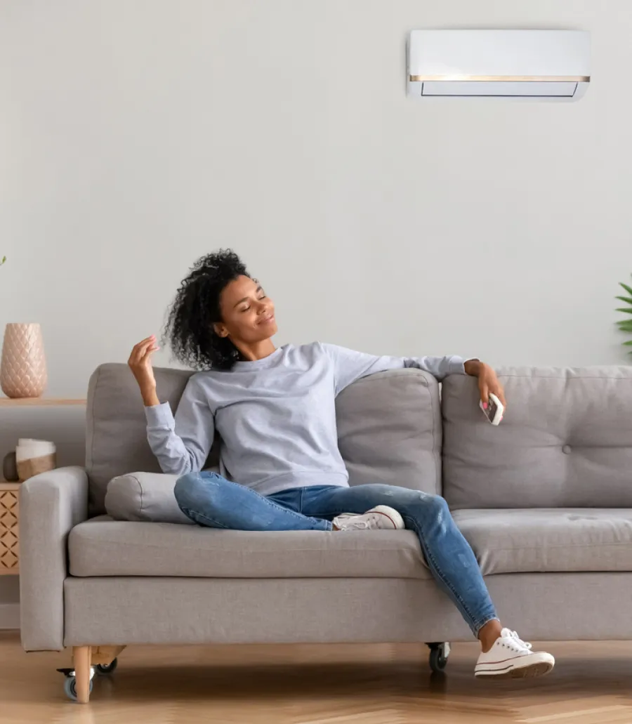 Woman relaxing on gray couch using remote to control wall-mounted air conditioner in modern living room