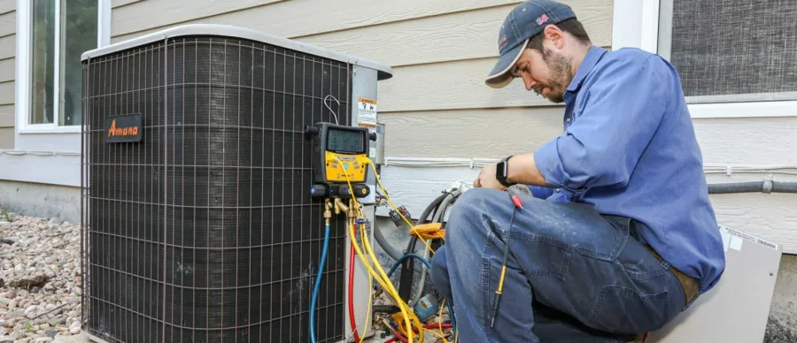 Technician in blue uniform repairing an outdoor Amana HVAC unit with tools and gauges near a house wall.
