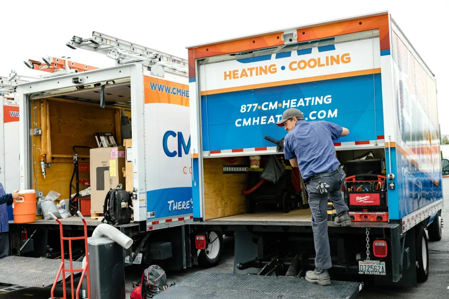 Technician loading tools into CM Heating and Cooling service trucks with ladders and equipment outdoors.