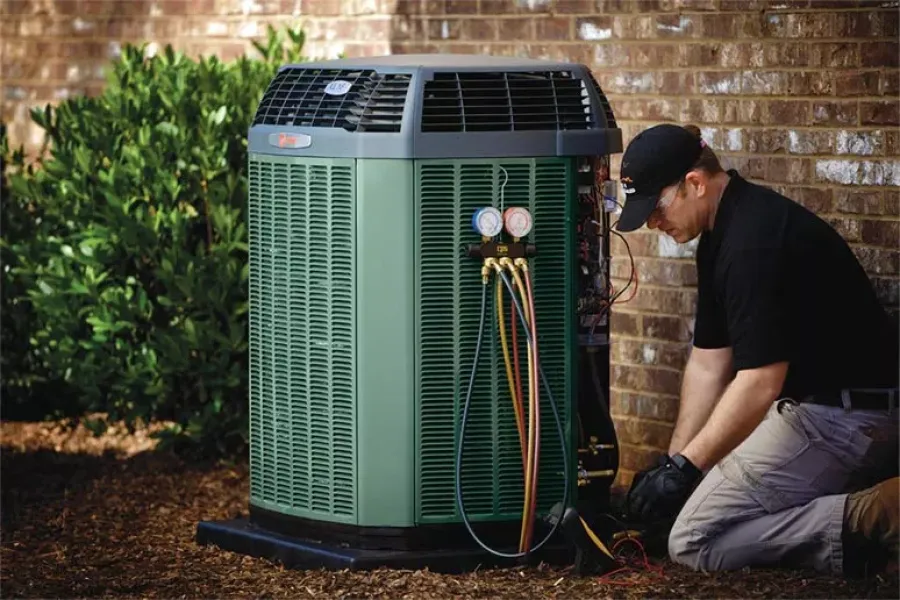 Technician servicing a green outdoor air conditioning unit attached to a brick wall with gauges and cables.