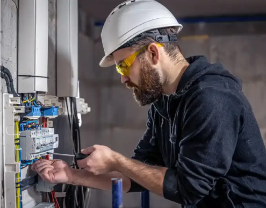 Electrician wearing safety helmet and glasses working on electrical panel with screwdriver indoors