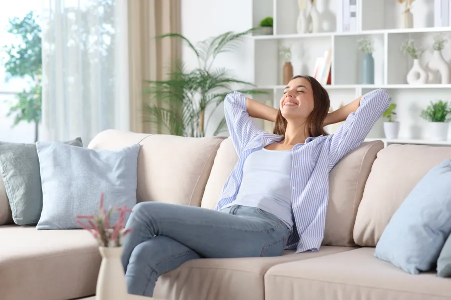 Woman relaxing on a gray sofa with a modern white air purifier in a bright living room with green plants.